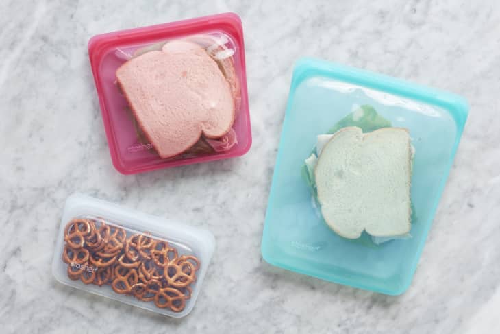 Sandwiches in pink and blue reusable silicone bags, with pretzels in a clear bag on a marble surface.
